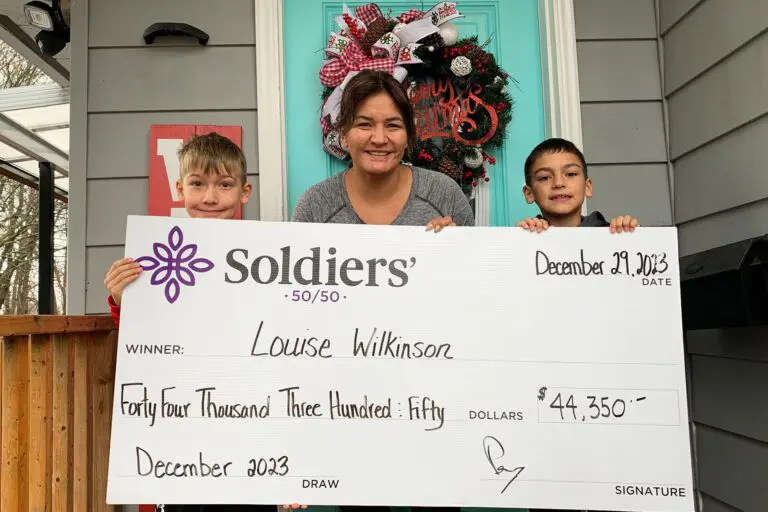 A women and her two young sons stand on a porch with a Christmas wreath on the door. They hold up a huge cheque for $44,350