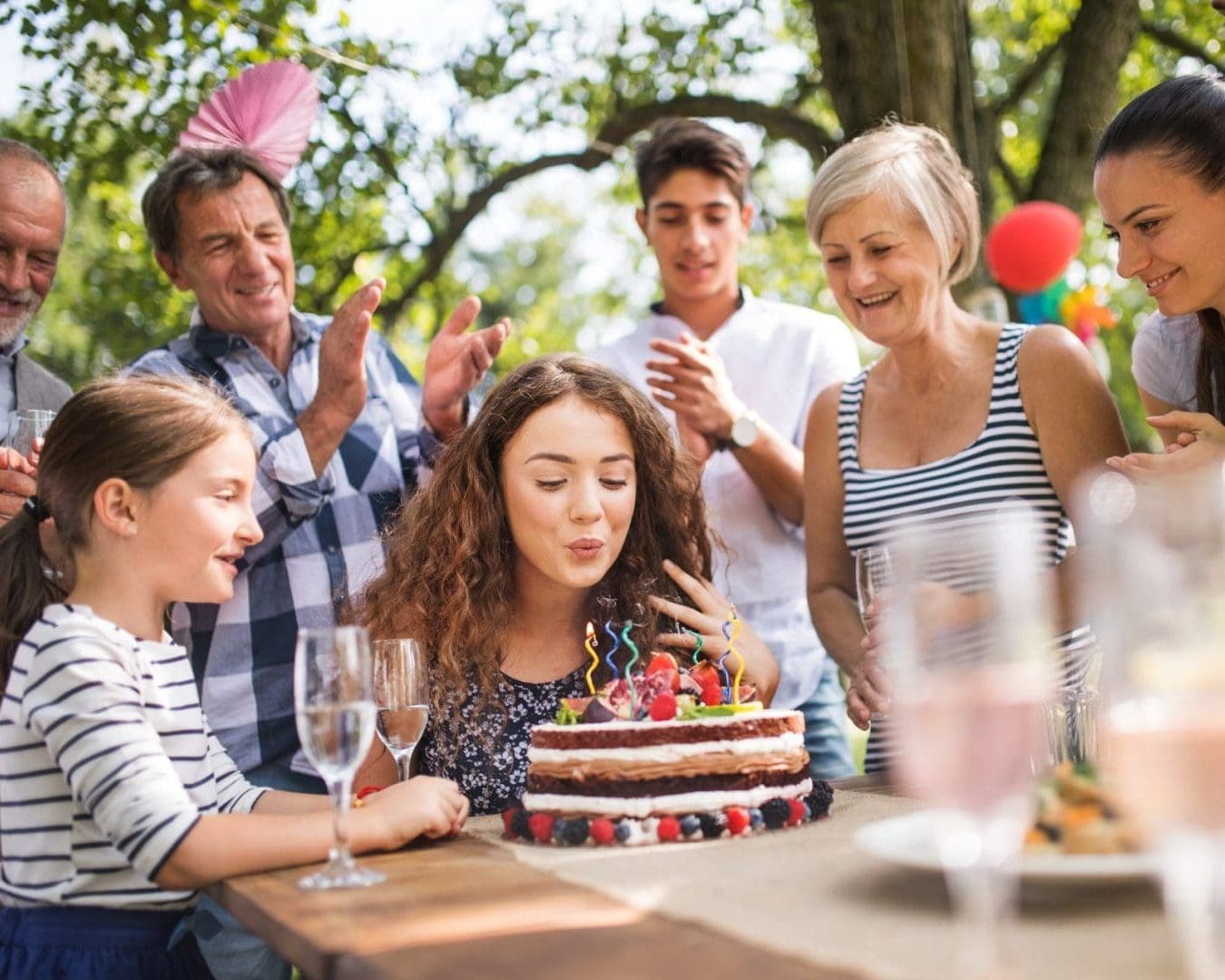 Youing girl with curly brown hair blows out a candle on a chocolate birthday cake while surrounded by family and friends