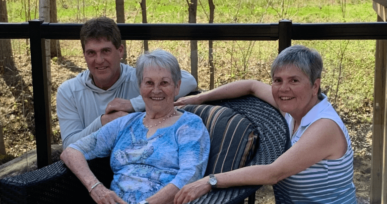 An elderly woman smiles in a chair with a middle aged man and woman smiling behind her