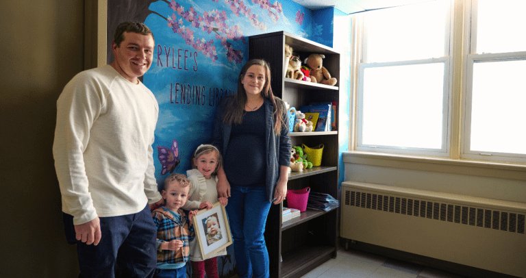 a man and a woman stand with their two small children in front of a blue wall. There is a mural with butterflies, flowers, and nature. Beside them is a book shelf with books for patients and their families to read during their stay.