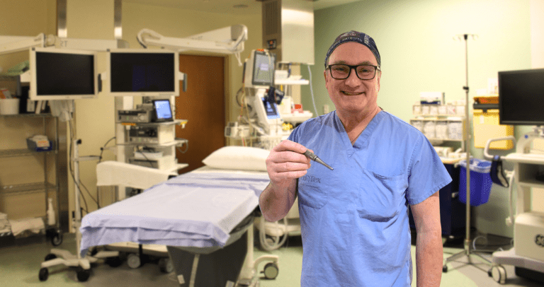 A middle aged white man wearing blue scrubs holds a new ear drill in an operating room.