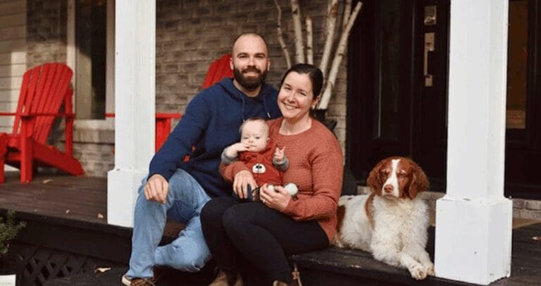 A white man with a brown beard and a white woman with brown hair sit out a porch, holding a baby boy with Down Syndrome. Beside them, a white and red dog lays on the deck.