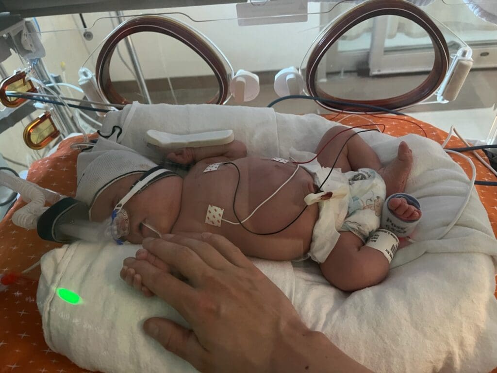 a small baby in a blue cap lays in a panda warmer at the NICU. His parent's hand can be seen reaching toward him
