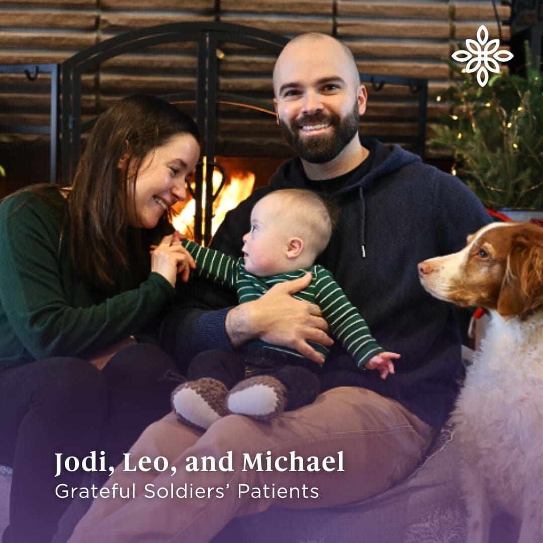 a white woman with brown hair sits beside a white man with a brown beard. There's a lit fireplace and Christmas tree behind them. The woman is leaning in to the infant sitting on the man's lap and a red and white dog sits beside them.