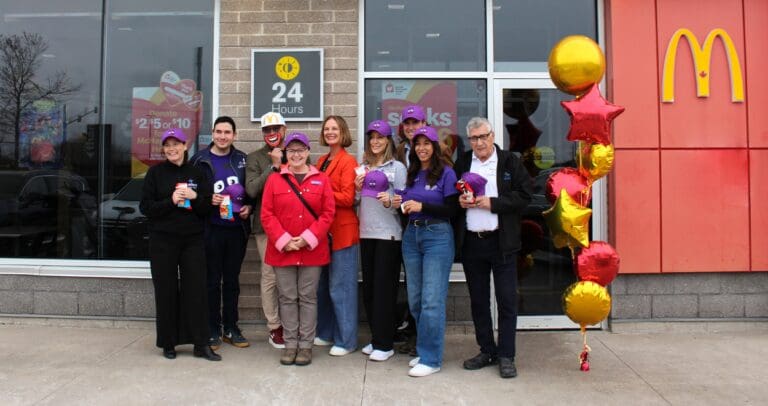 **Alt text:** Group of community members standing outside a McDonald’s restaurant holding purple hats, with a “24 Hours” sign and fundraising posters visible in the window, and red and gold star-shaped balloons displayed near the entrance.