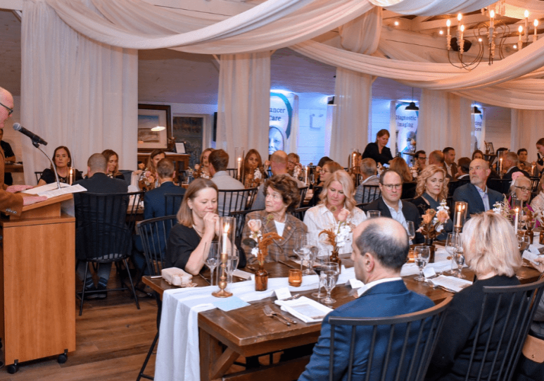 A man, Perry Esler, speaks in front of a crowd of people seated along two tables.