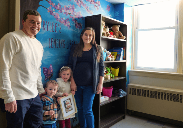 a man and a woman stand with their two small children in front of a blue wall. There is a mural with butterflies, flowers, and nature. Beside them is a book shelf with books for patients and their families to read during their stay.