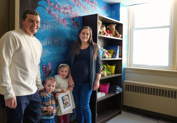 a man and a woman stand with their two small children in front of a blue wall. There is a mural with butterflies, flowers, and nature. Beside them is a book shelf with books for patients and their families to read during their stay.