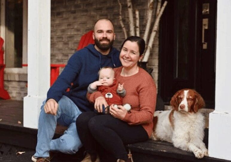 A white man with a brown beard and a white woman with brown hair sit out a porch, holding a baby boy with Down Syndrome. Beside them, a white and red dog lays on the deck.