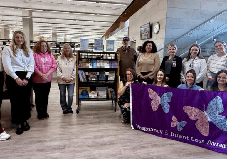 11 women and one man stand in a library beside a small bookshelf while holding up a purple flag with a pink and blue butterfly for pregnancy and infant loss awareness.