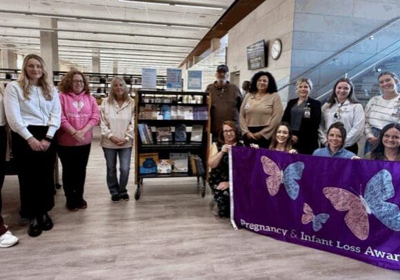 Little Loss Library 11 women and one man stand in a library beside a small bookshelf while holding up a purple flag with a pink and blue butterfly for pregnancy and infant loss awareness.