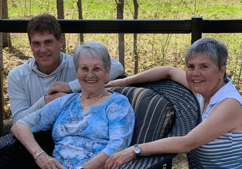 An elderly woman smiles in a chair with a middle aged man and woman smiling behind her