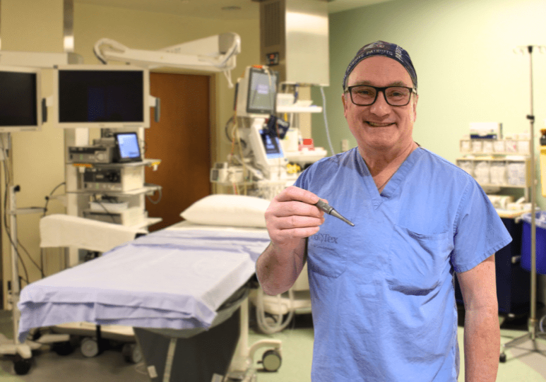 A middle aged white man wearing blue scrubs holds a new ear drill in an operating room.