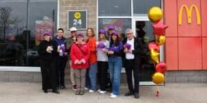 **Alt text:** Group of community members standing outside a McDonald’s restaurant holding purple hats, with a “24 Hours” sign and fundraising posters visible in the window, and red and gold star-shaped balloons displayed near the entrance.