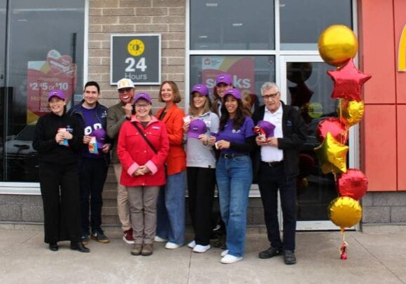 **Alt text:** Group of community members standing outside a McDonald’s restaurant holding purple hats, with a “24 Hours” sign and fundraising posters visible in the window, and red and gold star-shaped balloons displayed near the entrance.