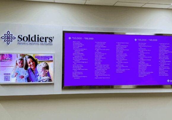A beige wall with a large digital screen on it. The screen is purple and has a list of donor names written in white. To the right, there is a "Soldiers'" logo and a screen with a doctor talking to two children.