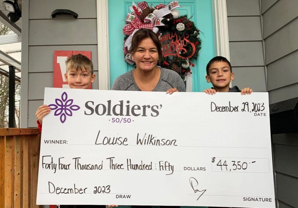 A women and her two young sons stand on a porch with a Christmas wreath on the door. They hold up a huge cheque for $44,350