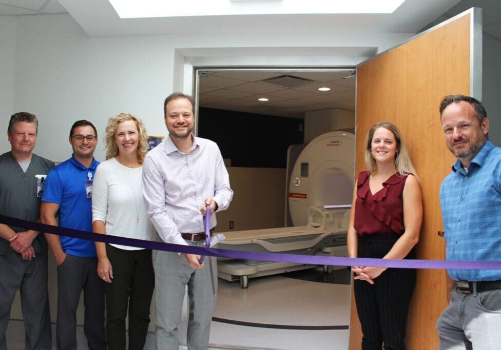 Four men and two women pose holding a large ribbon at the entrance to a door showing an MRI machine inside.